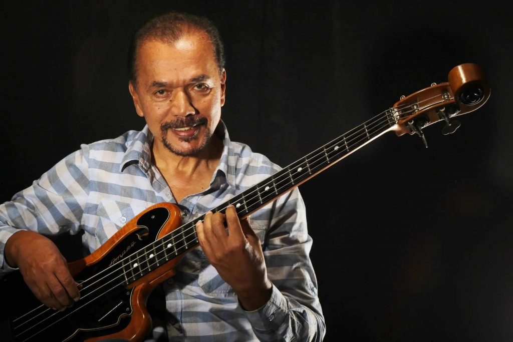 El músico y bajista de salsa Bobby Valentín sonríe a la cámara mientras sostiene su bajo, en una foto de estudio con fondo negro, previo a recibir su Doctorado Honoris Causa.