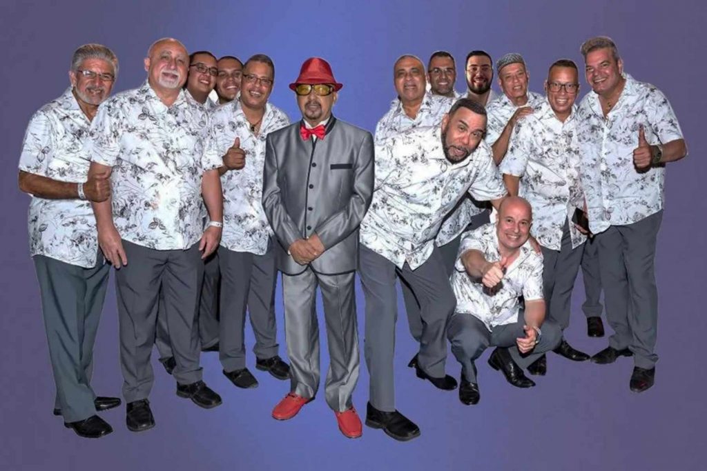 El pianista Papo Lucca y La Sonora Ponceña en una foto grupal. Papo Lucca destaca con un traje gris y sombrero rojo, como el líder de una de las orquestas de salsa más famosas de Puerto Rico.