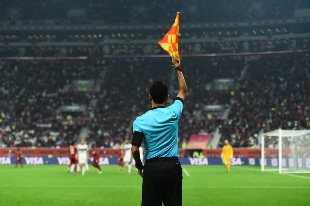 Árbitro asistente levantando la bandera para señalar un fuera de lugar durante un partido, la señal visual que indica la infracción a la regla explicada en la nota.