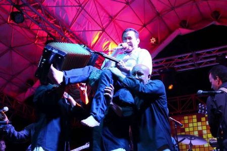 Alfredo Gutiérrez ejecutando el acordeón con los pies mientras es cargado por su equipo en una presentación en vivo.