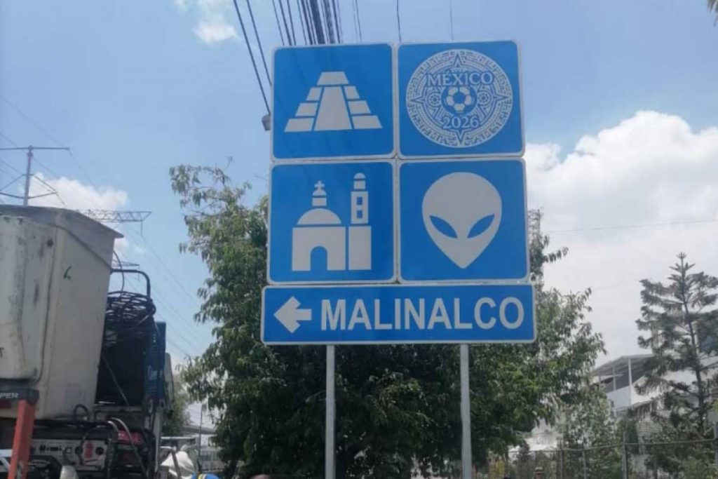 Letrero azul hacia Metepec ubicado junto a la estructura del Tren Interurbano México-Toluca, con iconos de artesanías (barro), una iglesia, un cóctel y el logo de México 2026.