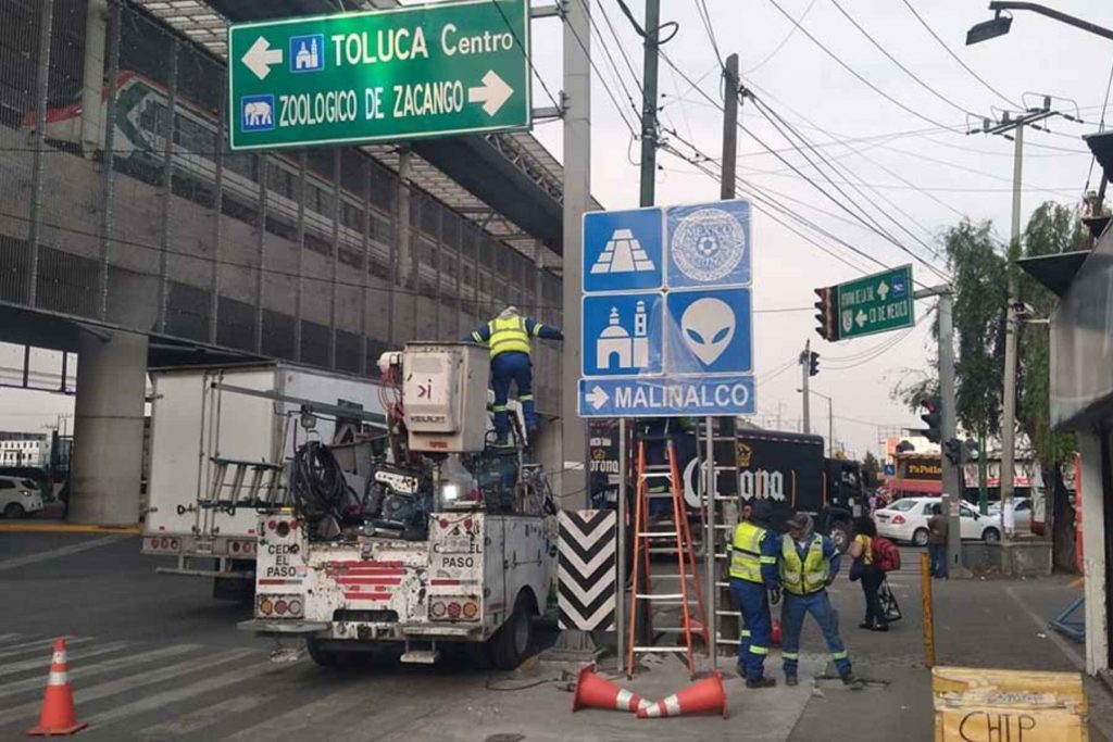 Trabajadores con chalecos de seguridad instalando nuevos señalamientos viales de Toluca Centro, el Zoológico de Zacango y Malinalco en una avenida transitada.