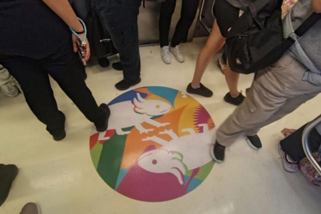 Calcomanía circular con fondo multicolor y figuras de dos ajolotes blancos en el piso del Metro CDMX rodeada por los pies de los usuarios.